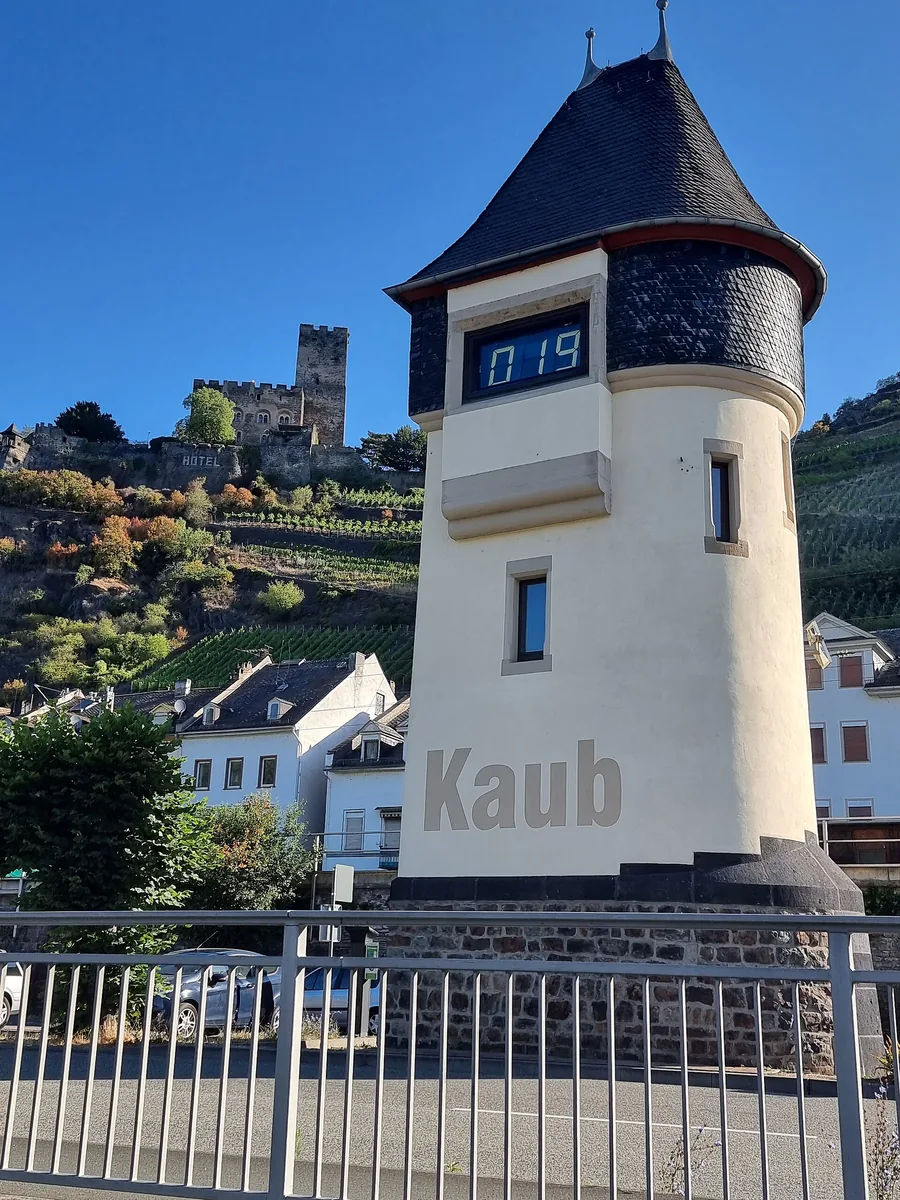 A white round tower with a pointed slate roof and a digital display showing the figures 019, with the name Kaub on the wall. Behind the tower, a slope with vines and a castle under a clear blue sky.