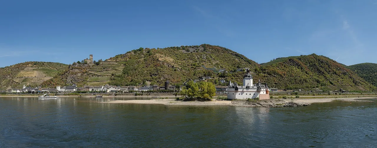 Panorama of the Rhine with the white Pfalzgrafenstein Castle on an exposed sandbank; behind it the small town of Kaub on the bank and wooded hills with vineyards under a blue sky.