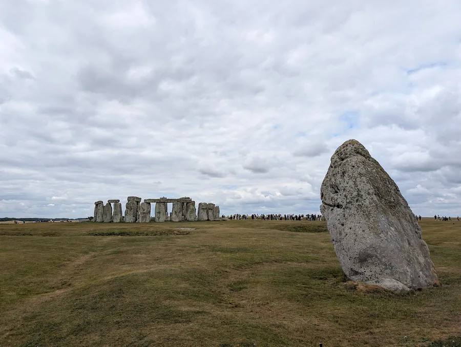 A large, leaning rough stone on the right in the foreground; on the left in the distance the stone circle with a row of small visitors under a cloudy sky.