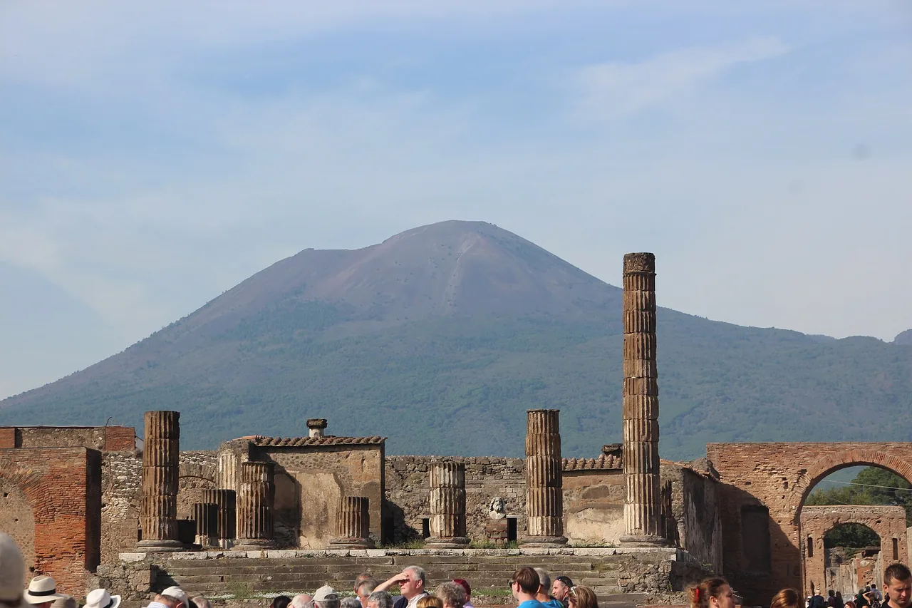 Ruins with free-standing columns and brick arches in the foreground, behind them the broad cone of Vesuvius and a pale blue sky.