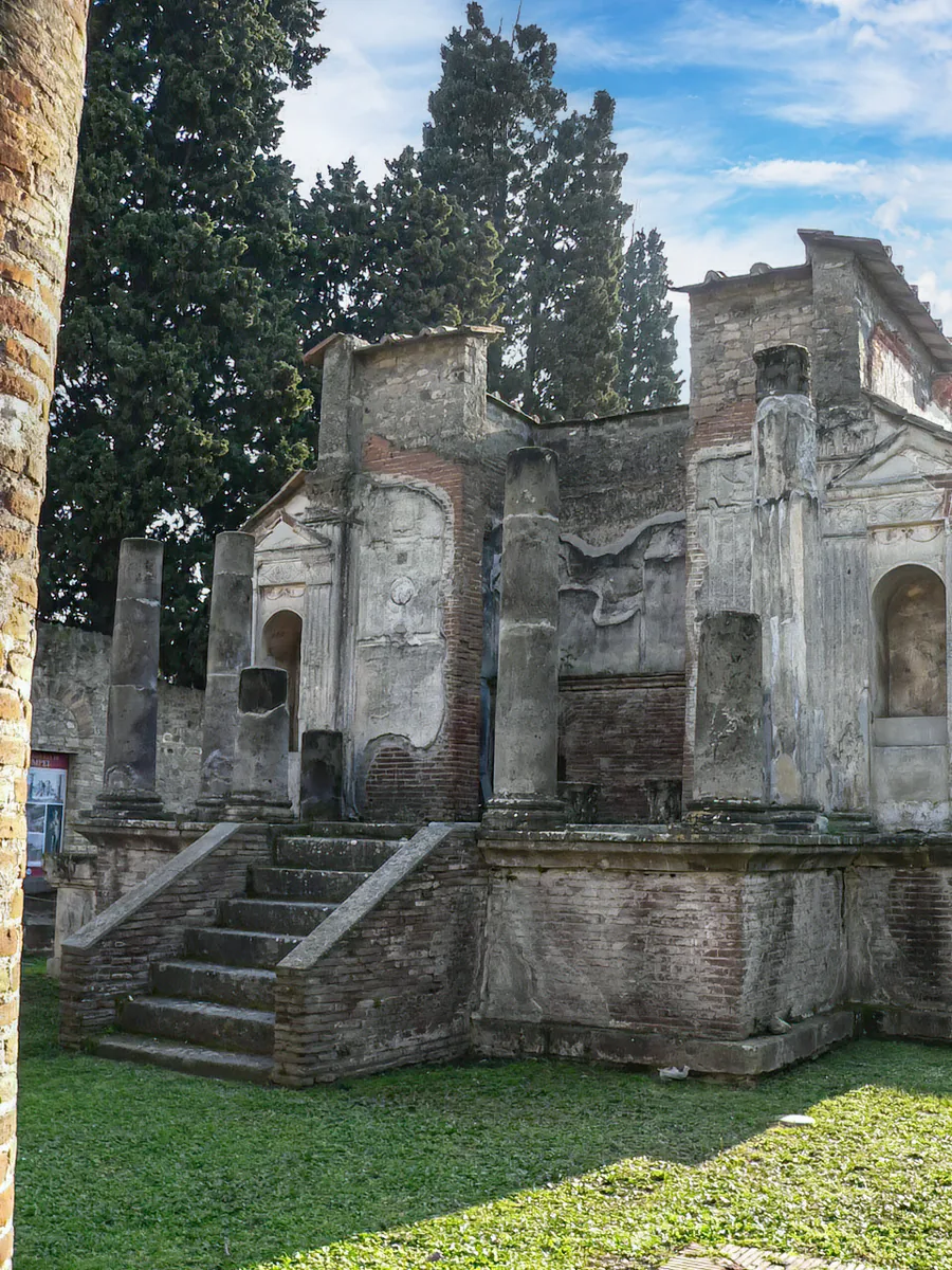 The ruin of a small temple on a stone podium with steps, broken columns and walls with niches, in front of tall dark cypresses and a blue sky.