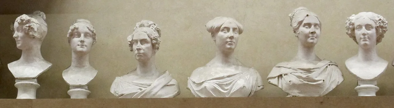 Six white plaster busts of women with different hairstyles stand side by side on a wooden shelf against a light wall.