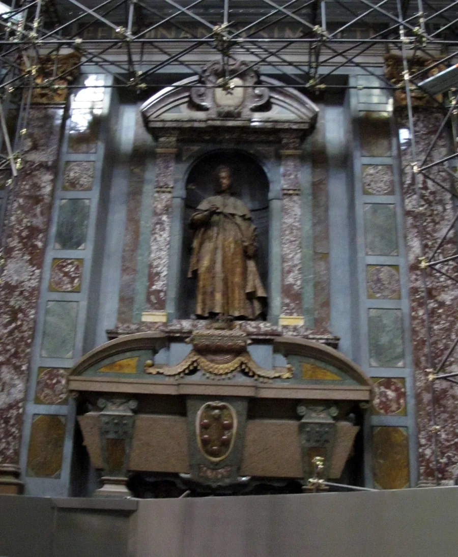 A dark bronze standing statue in a niche above a marble funerary monument, surrounded by coloured marble walls and metal scaffolding tubes.