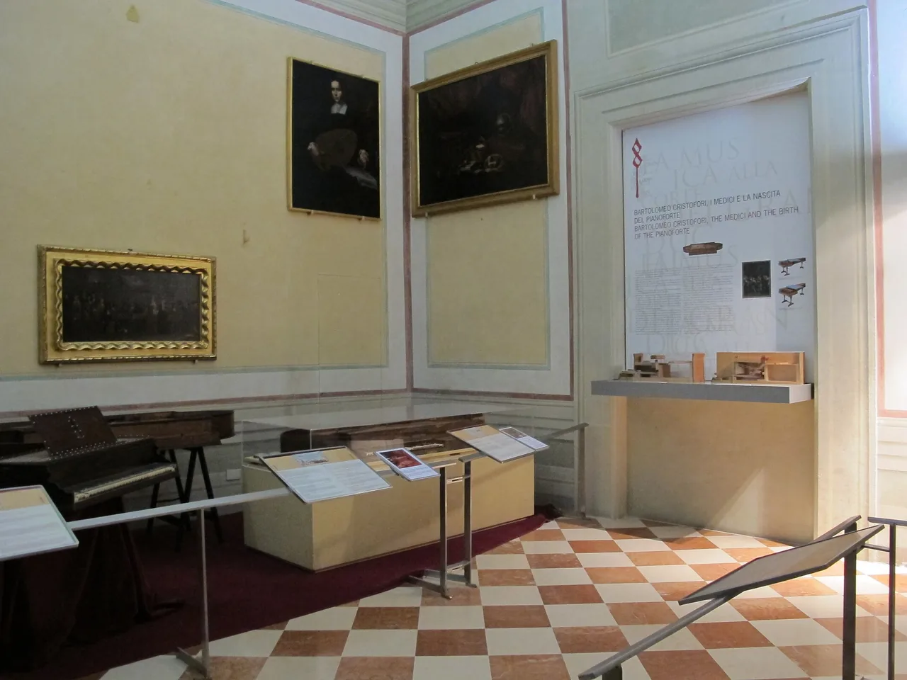 A museum room with a chequered floor, a display case with a keyboard instrument, paintings on yellow walls and an information panel about Cristofori.